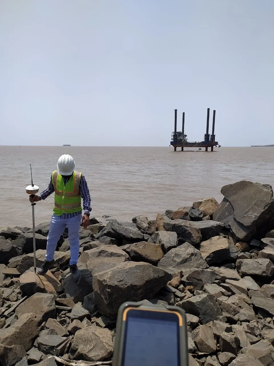 A surveyor with a GNSS pole and controller on a rocky shore